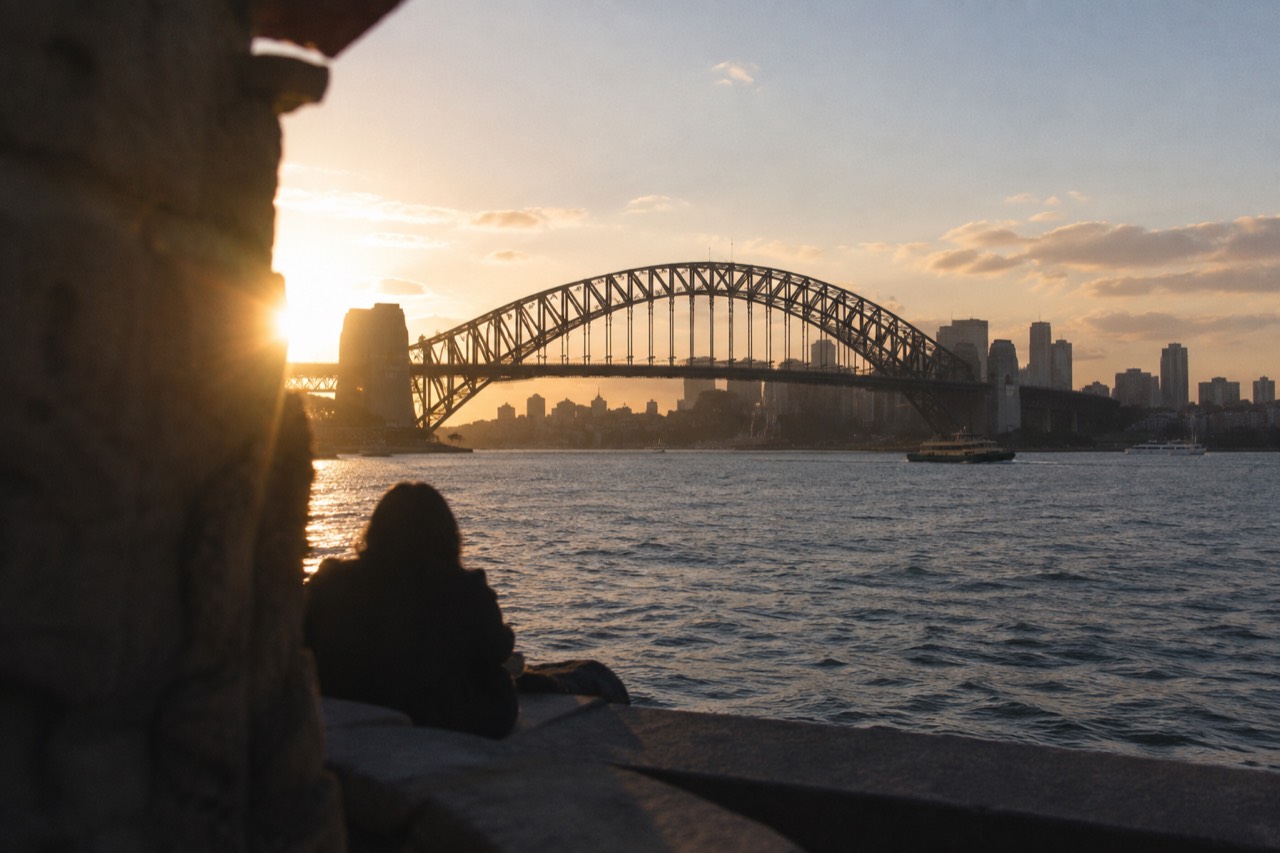 The steel arch of the Sydney Harbour Bridge rising above the water at dusk, with the city skyline behind it