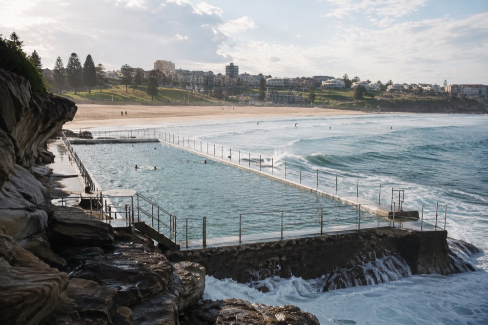 A long ocean pool set into rocks beside a curved beach with parkland behind it