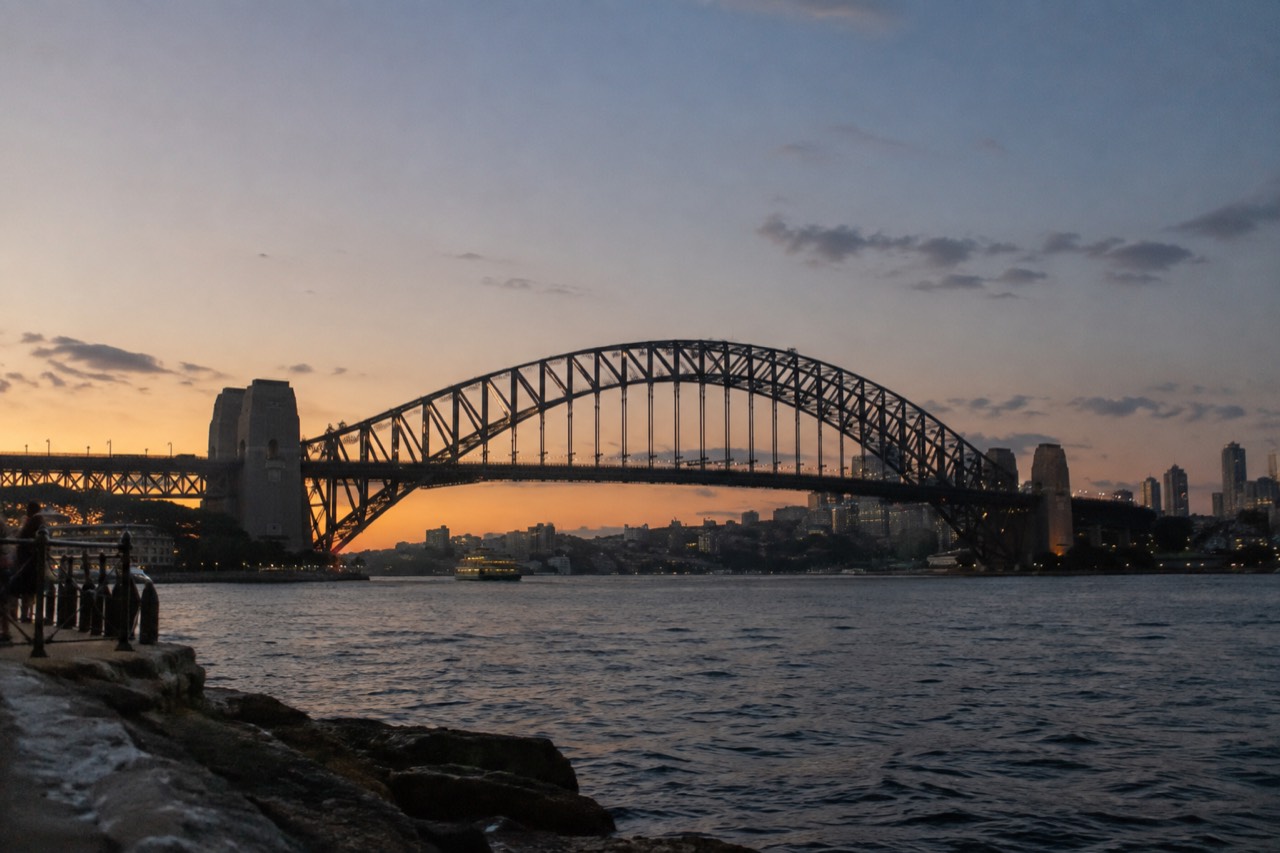 The steel arch of the Sydney Harbour Bridge rising above the water at dusk with the city skyline behind it