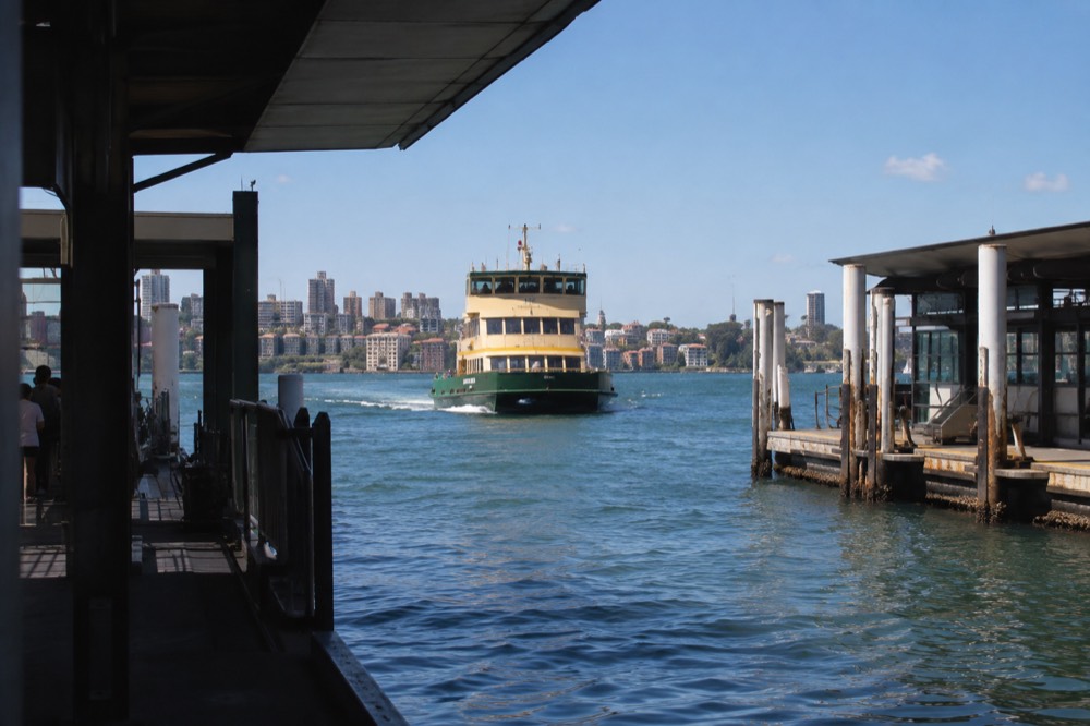 A Sydney ferry crossing the harbour toward Manly with ferry wharves along the shore