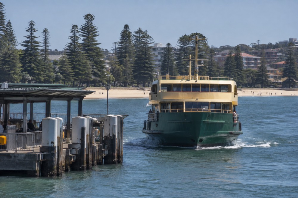 A green and yellow ferry approaching a wharf with a sandy beach and Norfolk Island pines behind it