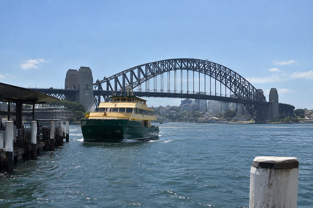 A green and yellow Sydney ferry approaching a wharf with the bridge rising above the water in the background