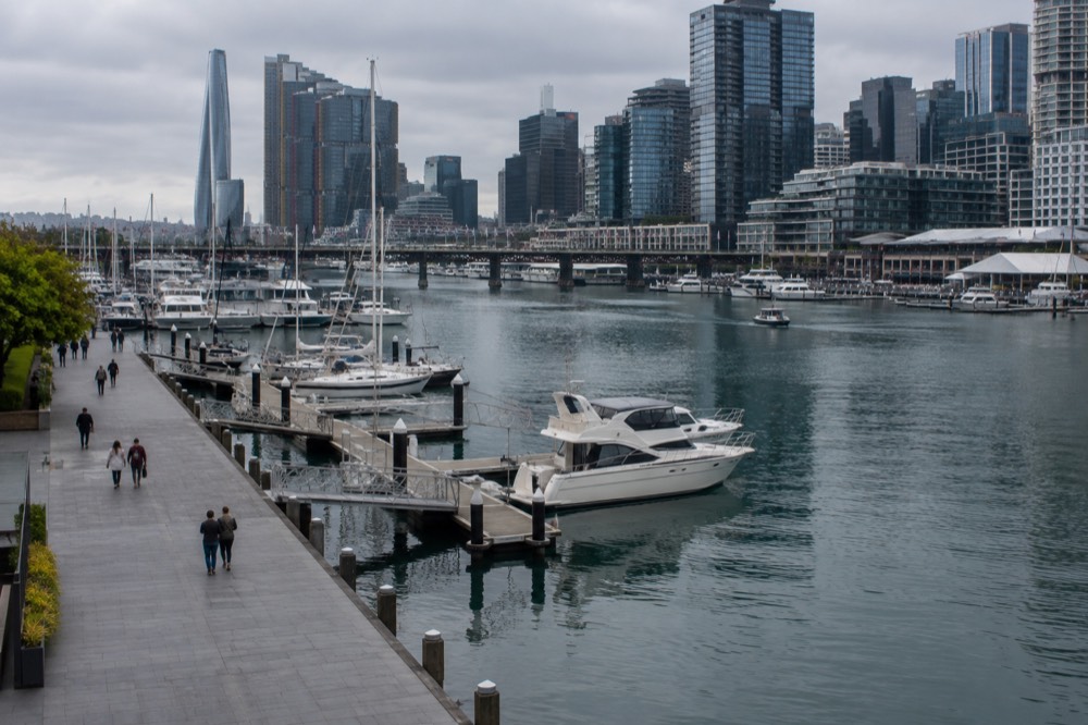 A level paved waterfront walkway along Darling Harbour with moored boats and high-rise buildings behind them