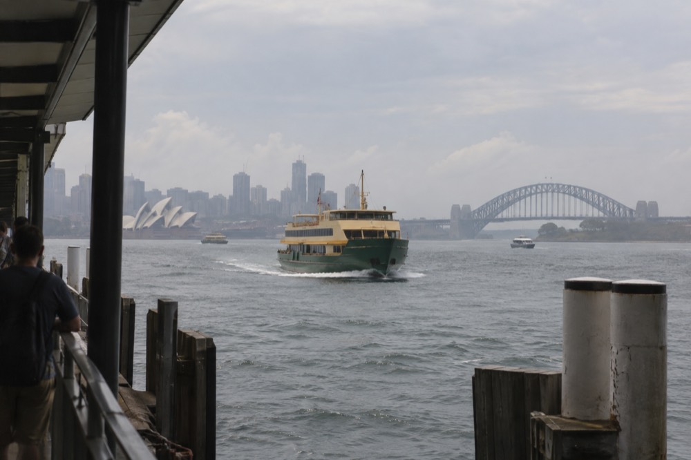 A green-and-yellow ferry crossing grey harbour water toward a city skyline under cloud
