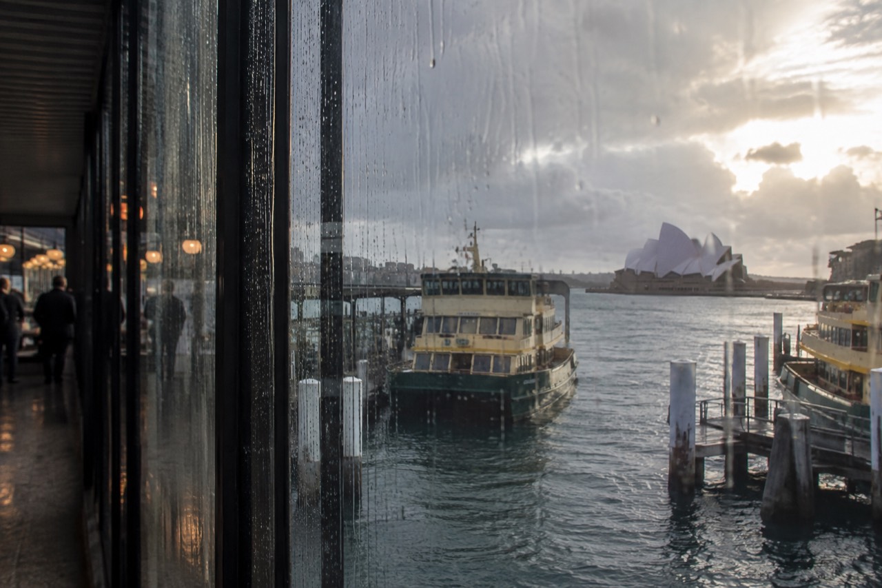 Rain streaking the glass of a harbour-side building with grey water and moored ferry boats beyond
