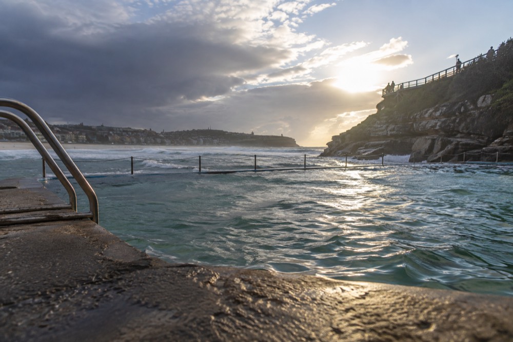 An ocean pool at the edge of a beach with a headland path beyond it