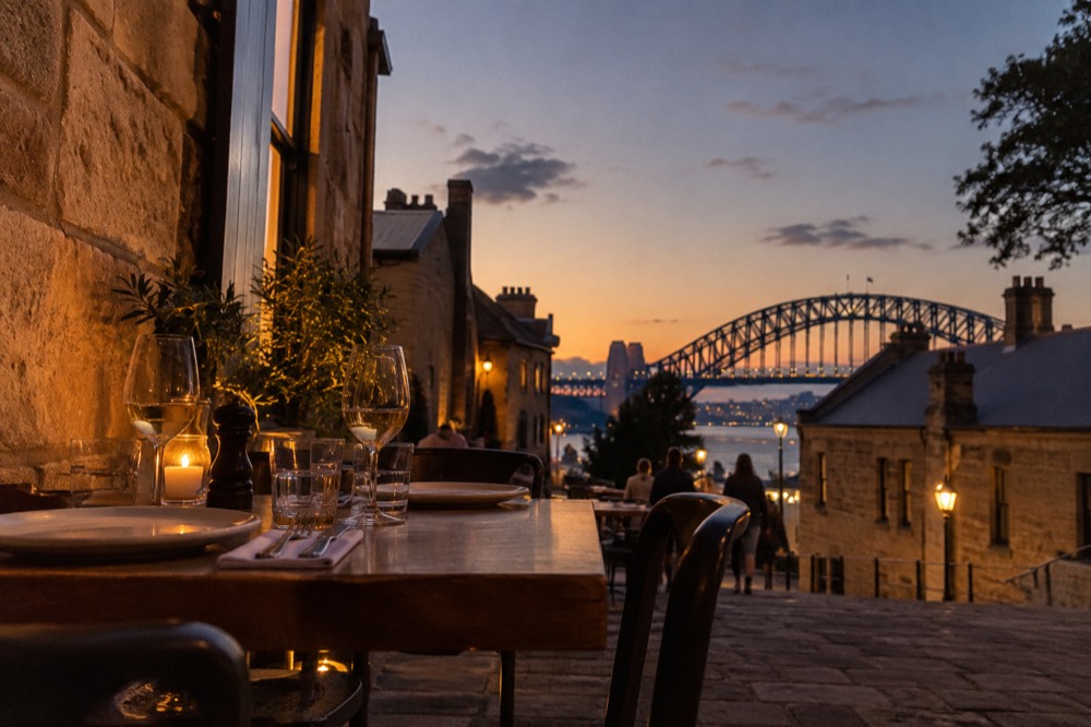 An evening table set beside a sandstone wall in The Rocks with the Harbour Bridge visible above the rooftops