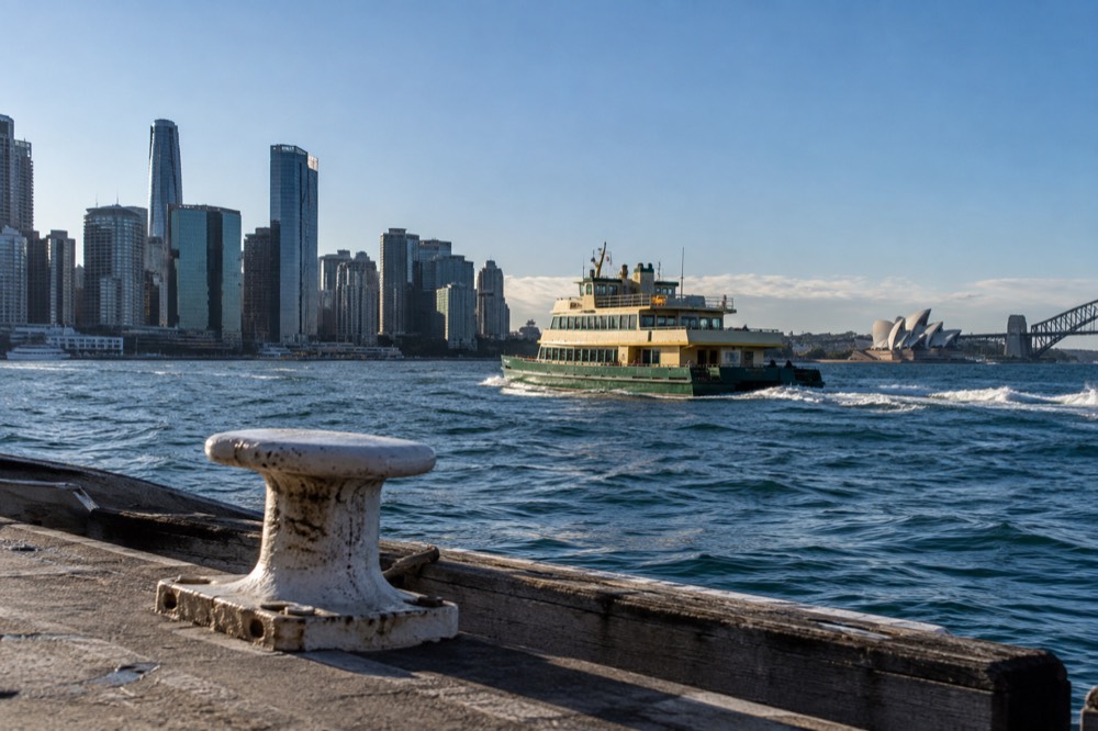 A green and yellow public ferry crossing Sydney Harbour towards Manly with the skyline behind it