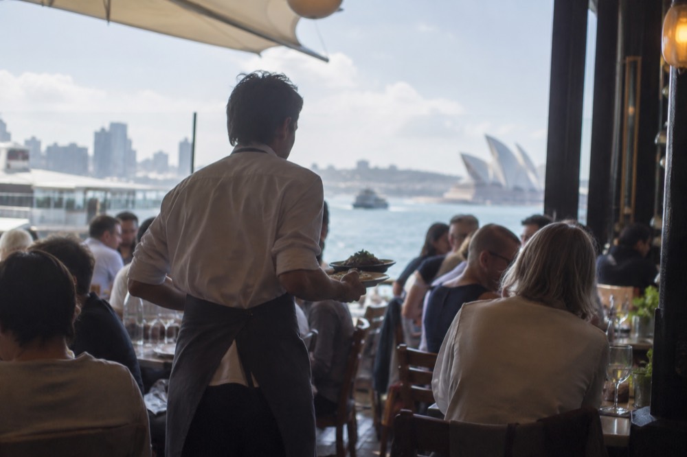 A waiter carrying plates through a busy narrow Sydney restaurant dining room
