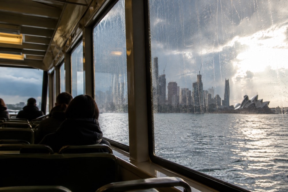 A covered ferry cabin on Sydney Harbour with rain streaking the windows and the skyline behind