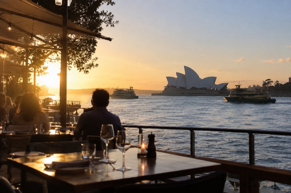 Harbourside restaurant tables at dusk overlooking Sydney Opera House and harbour ferries
