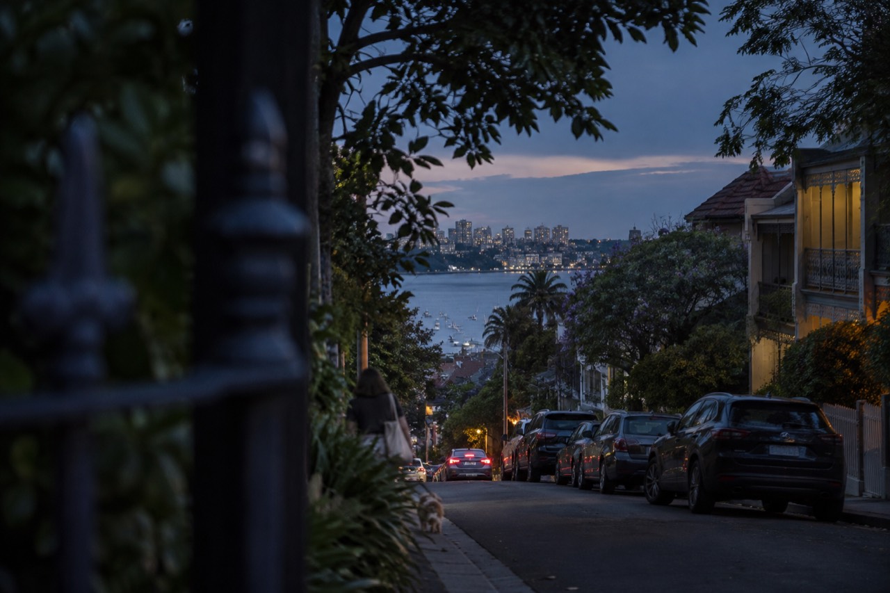 A leafy street of terrace houses sloping down toward a calm harbour bay in Glebe, Sydney