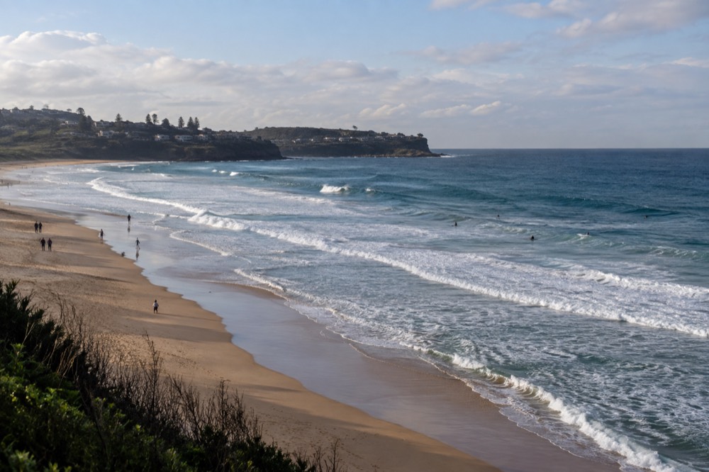 A wide sandy beach with rolling surf and a headland in the distance under morning light