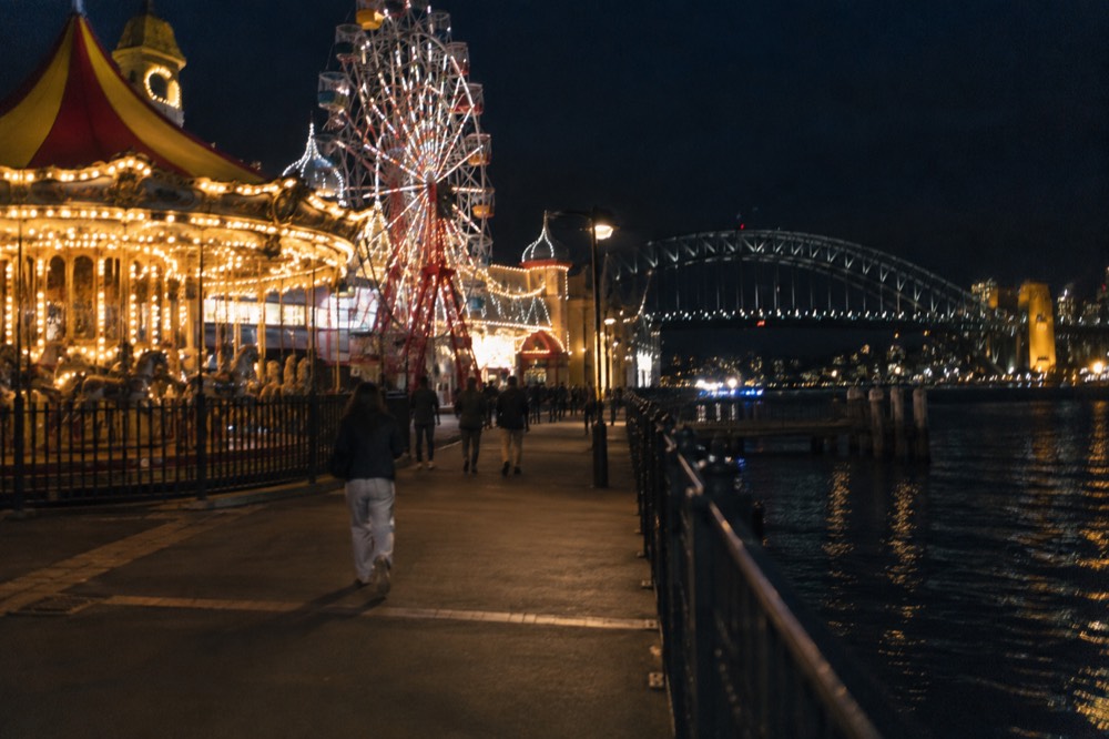 An amusement park lit up at night with a harbour bridge behind it