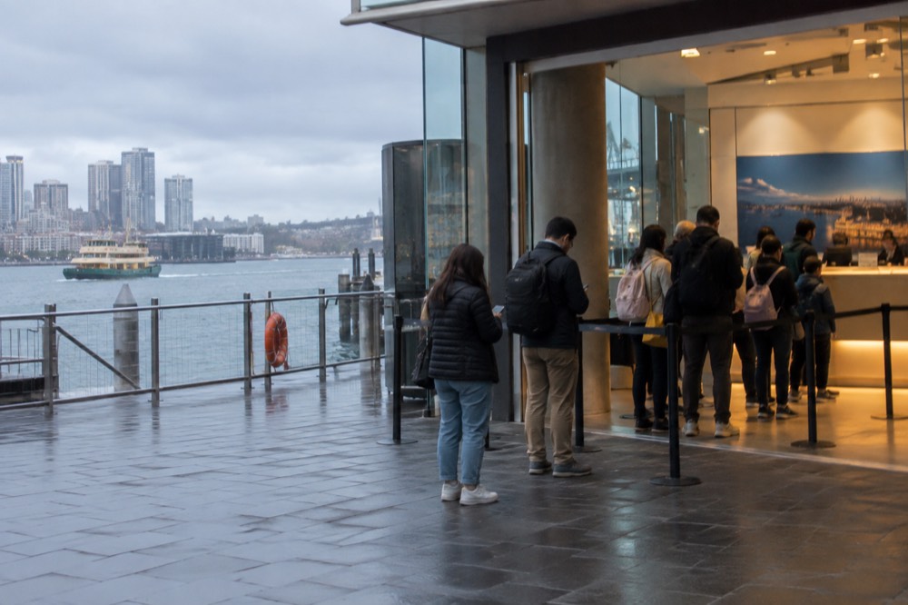 A ticket counter with a queue of visitors holding phones in a brightly lit attraction lobby