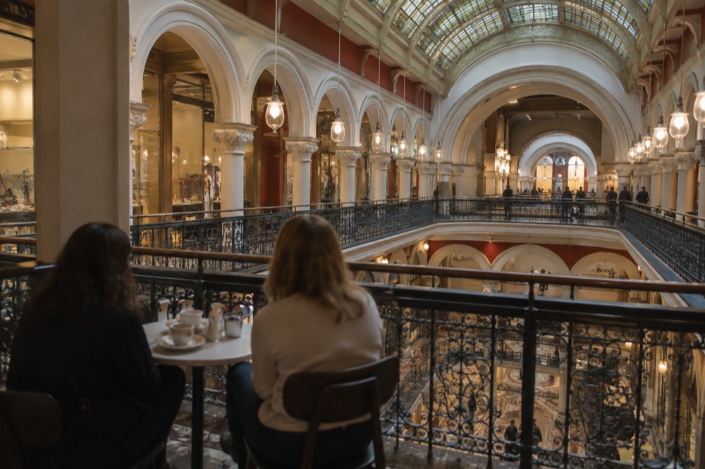 Two shoppers resting at a cafe table on an upper gallery overlooking an arcade atrium