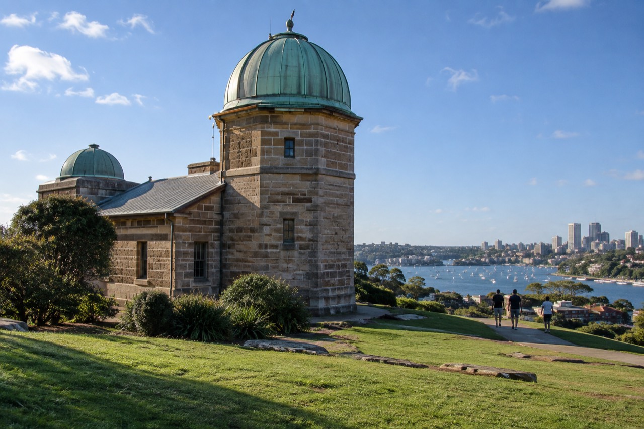 A historic stone observatory building with a domed tower on a green hill overlooking Sydney Harbour