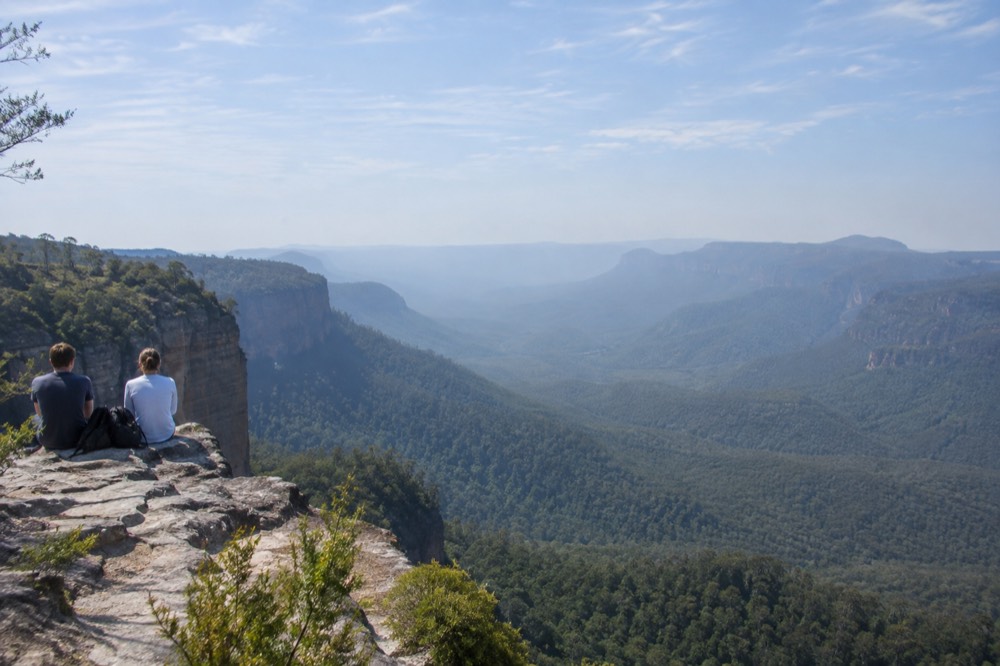 A wide valley of eucalypt forest filling with morning haze below sandstone cliffs