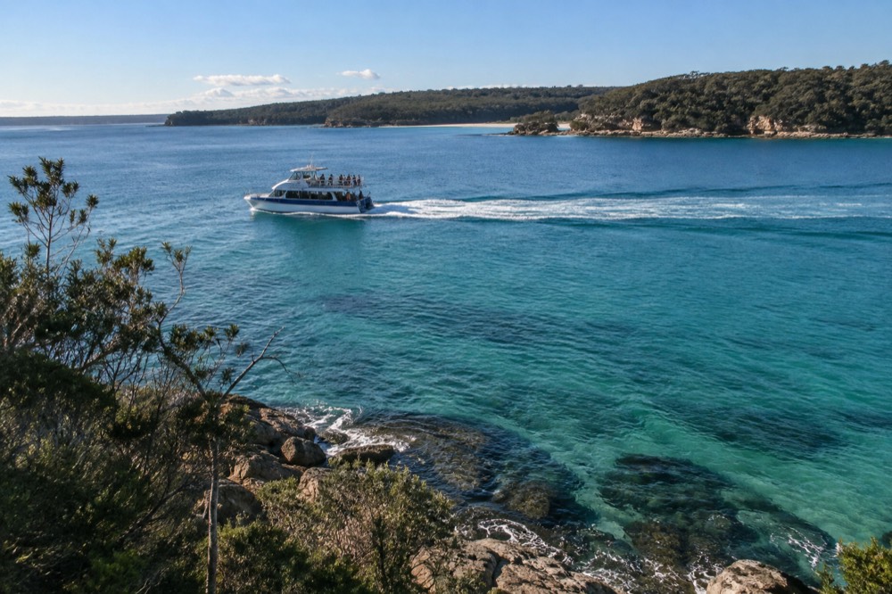 A tour boat cutting across clear turquoise water with forested headlands on the horizon