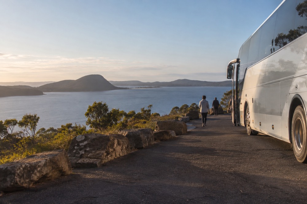 A coach parked beside a coastal road turnout with a bay visible below