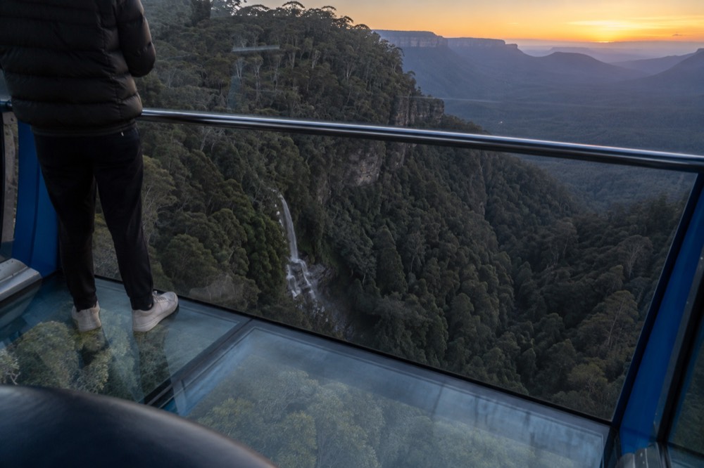 Glass-floored cable car moving above a forested gorge with a waterfall below