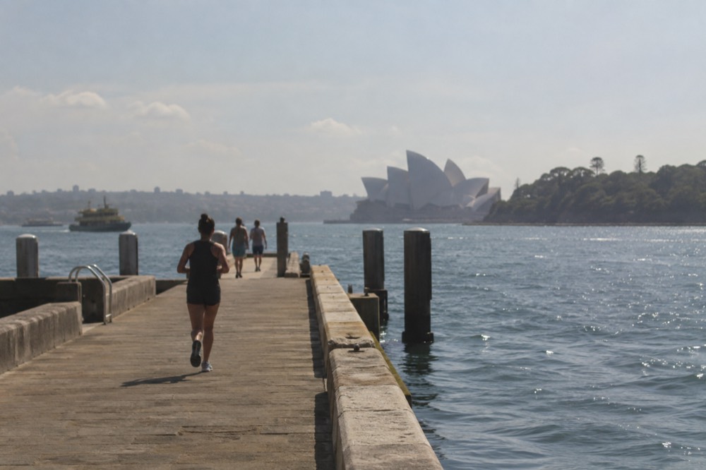 A stone jetty extending into harbour water with the Opera House visible behind a forested headland