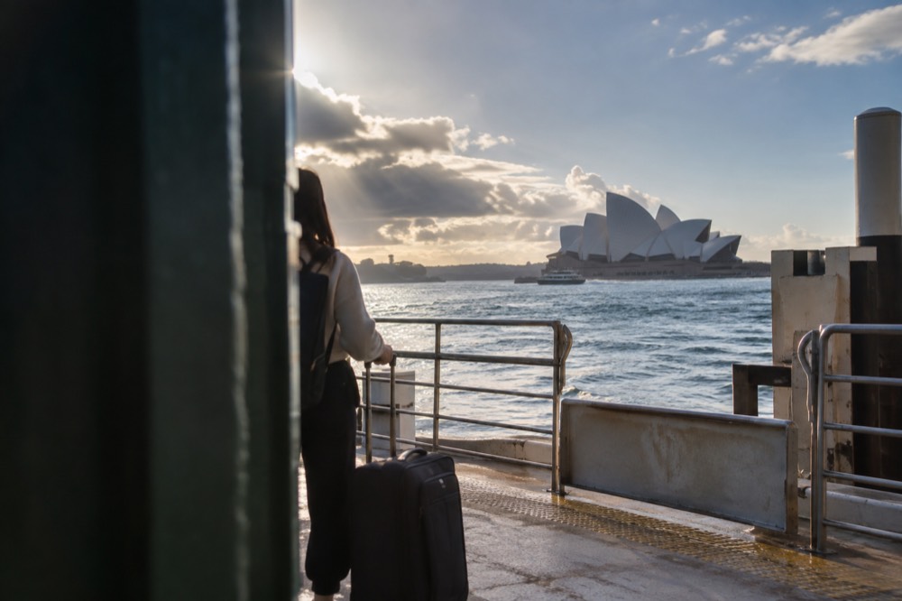A traveller standing with a suitcase on a ferry wharf with the Opera House in the background