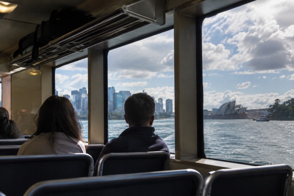 A ferry interior with a luggage rack and passengers seated near a window looking out at the harbour