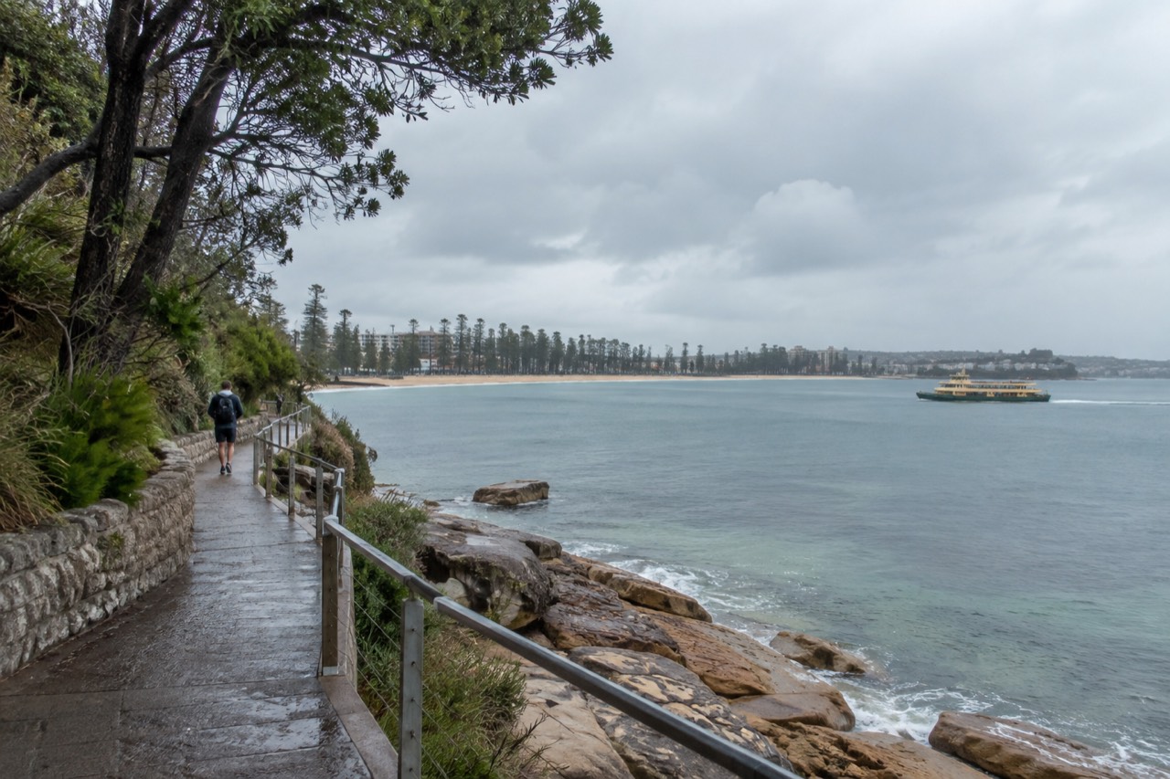 Sandstone headland path above calm harbour water with ferry in the distance