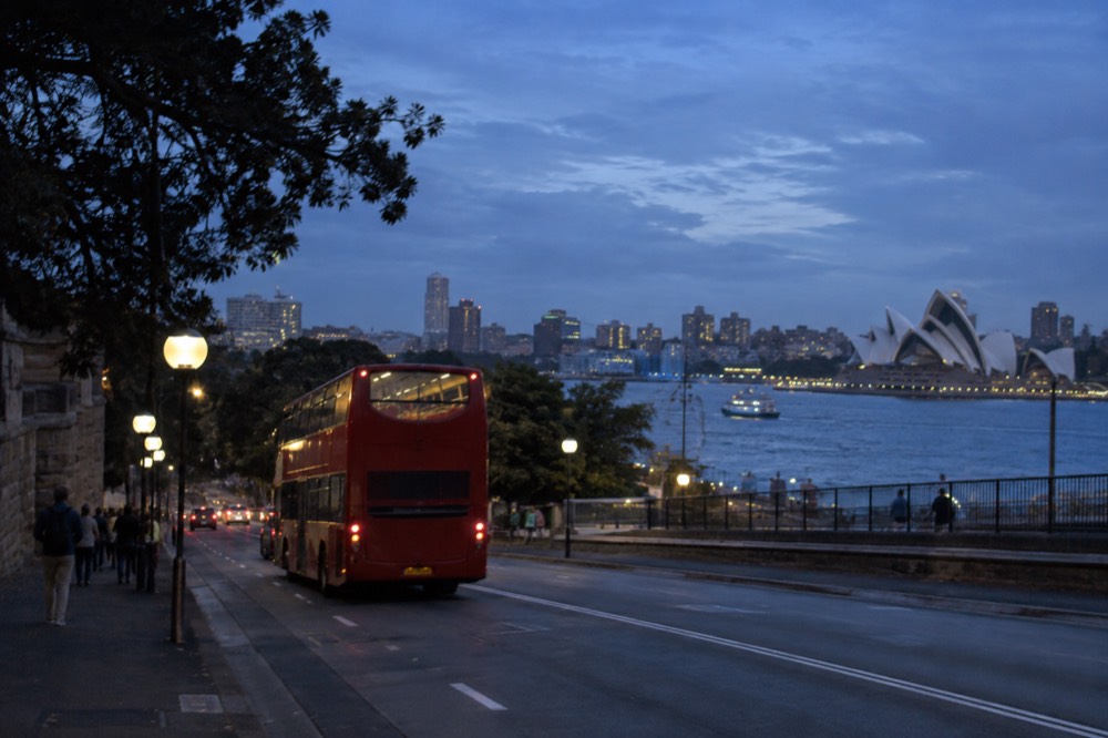 A red double decker sightseeing bus on a Sydney city street with the harbour in the distance