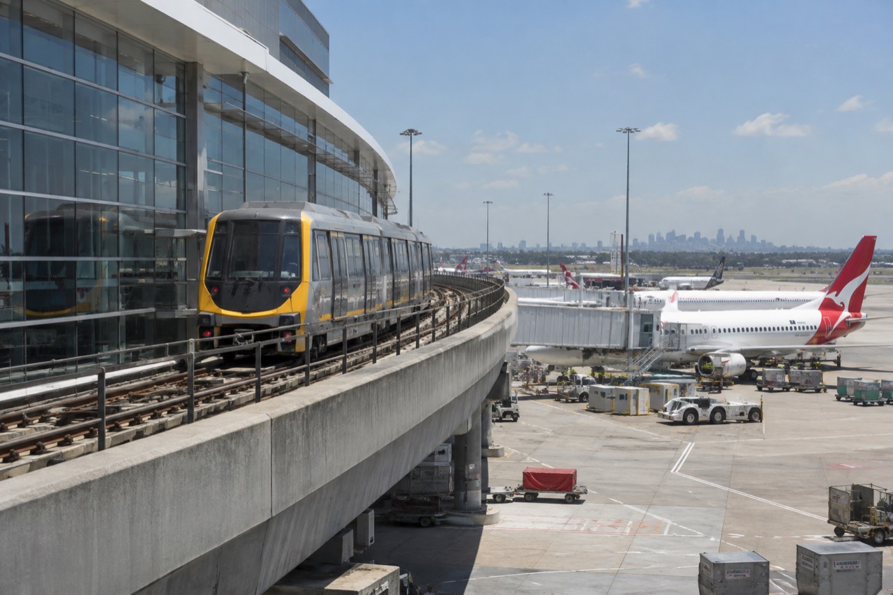 A train on the airport line beside a Sydney Airport terminal building under a clear sky