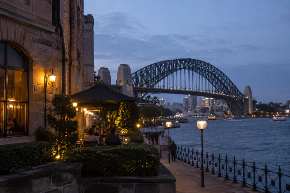 A heritage sandstone pub facade in The Rocks lit by evening lamps with the Harbour Bridge arch overhead