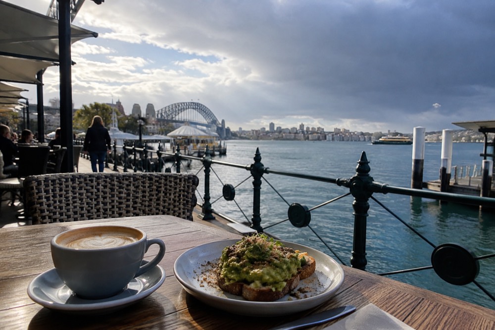 A flat white coffee in a ceramic cup beside a plate of avocado on toast on a café table
