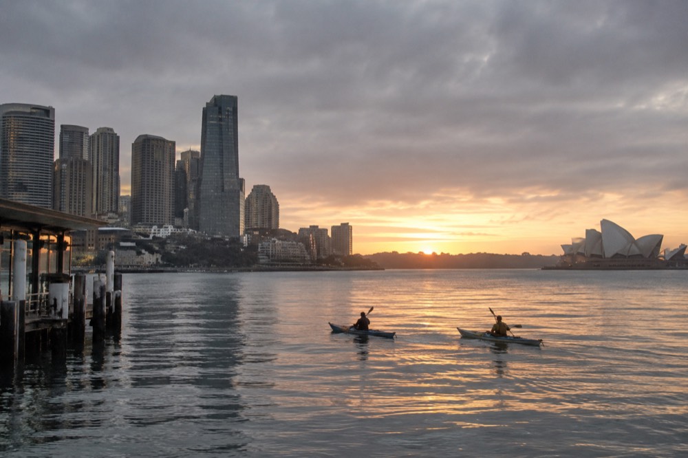 Two kayaks on glassy harbour water at sunrise with the city skyline behind
