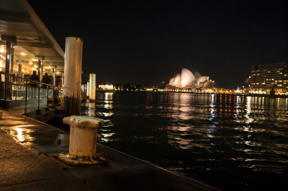 City lights reflecting on harbour water at night with the illuminated Opera House in the distance
