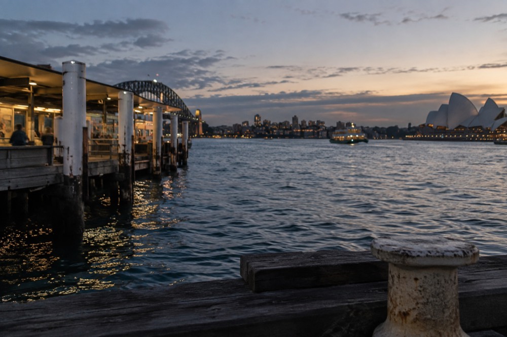 Camera on tripod overlooking harbour water at dusk with city lights beginning