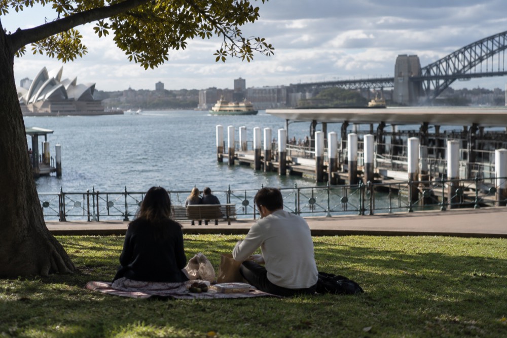 A couple unpacking food from a paper bag onto a blanket on a harbourfront lawn