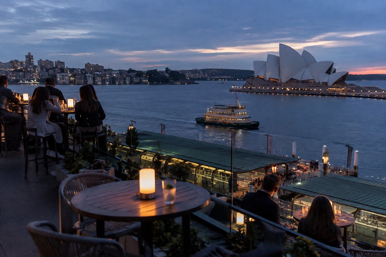 Rooftop bar tables overlooking Sydney Harbour at dusk with the Opera House in the distance