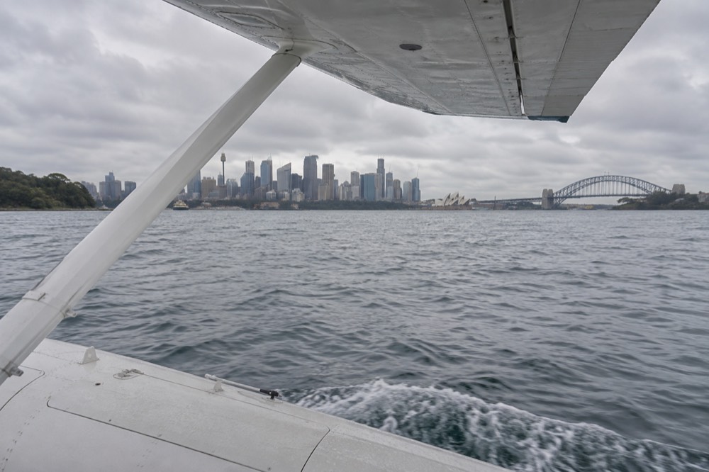 Seaplane wing above harbour water with the distant city skyline ahead