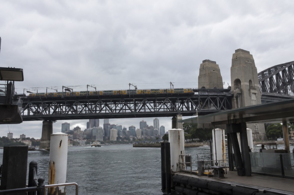 A Sydney commuter train crossing a steel truss bridge over the harbour with city towers behind it