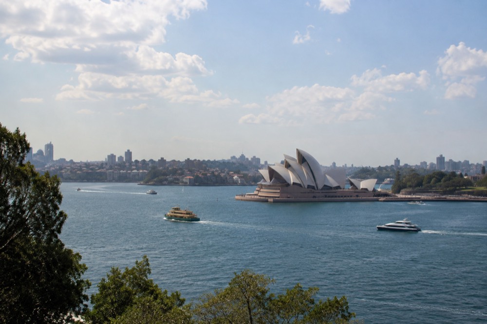 Sydney's harbour skyline under a wide sky, with the Opera House white sails against scattered clouds above blue water.