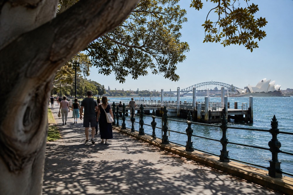 A wide garden path along a harbour shoreline with large trees and a distant bridge