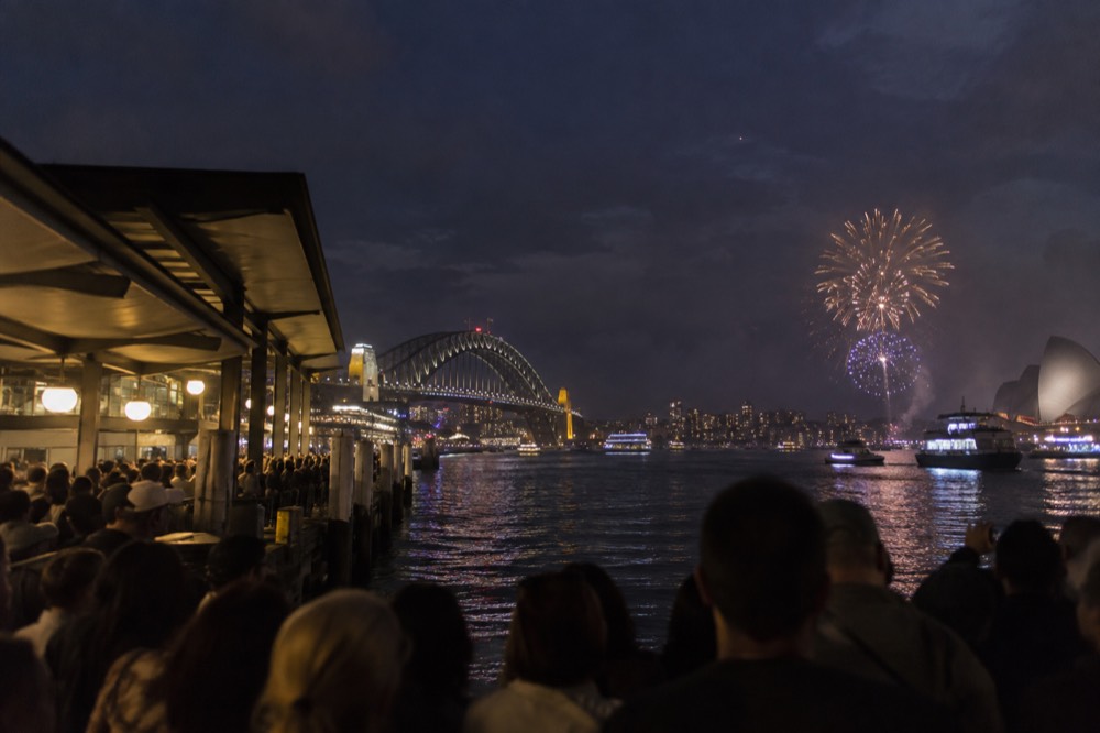 Crowds gathered along a harbour foreshore at dusk waiting for fireworks