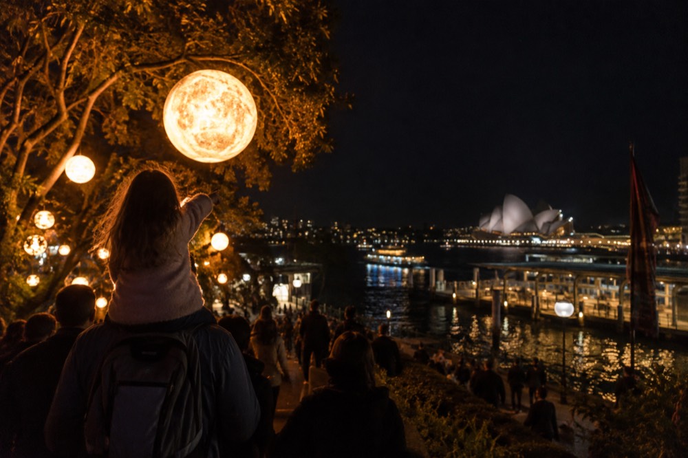 A child on a parent's shoulders pointing at a large glowing light installation in a park at night