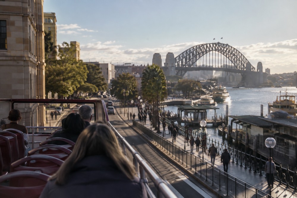 An open-top tour bus on a Sydney street with the Harbour Bridge visible ahead