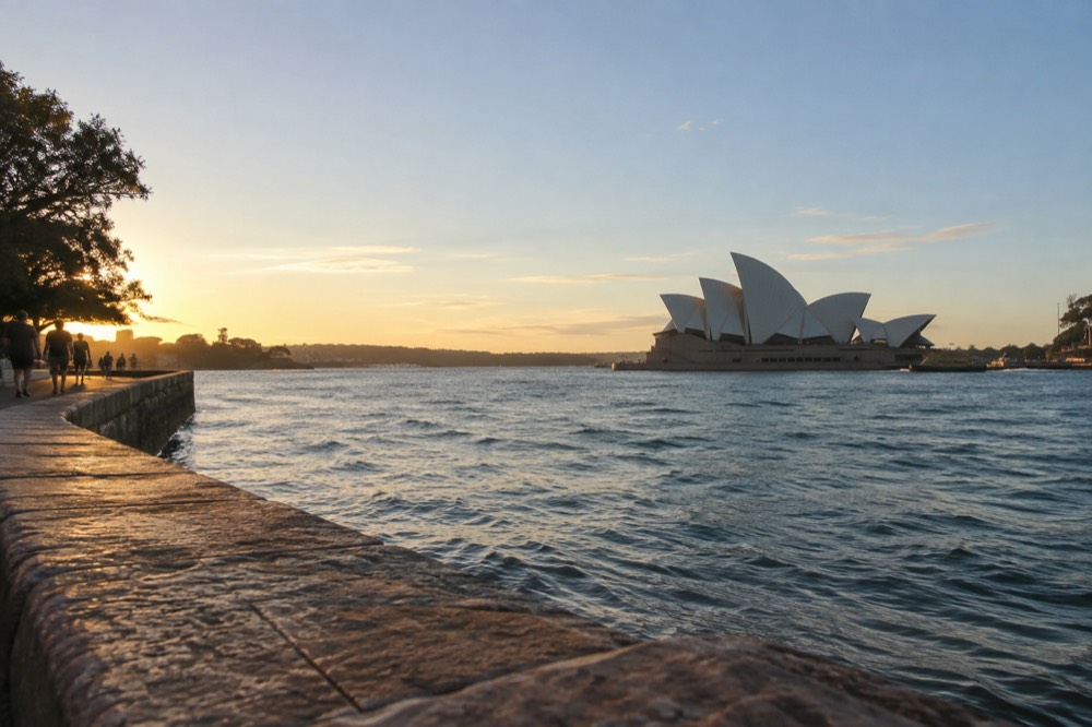 White sails of an opera house seen across a quay from a sandstone promenade